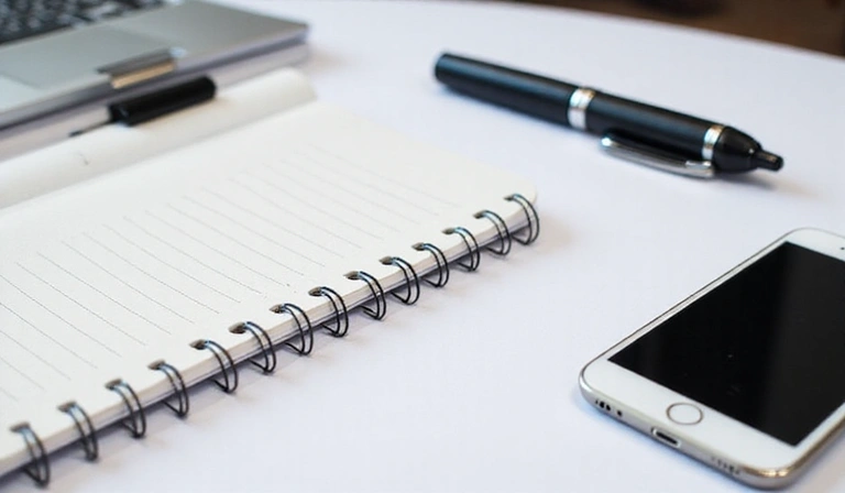 Ein Smartphone, ein Notizbuch und ein Stift liegen auf einem Schreibtisch, symbolisierend Kommunikationsmittel.